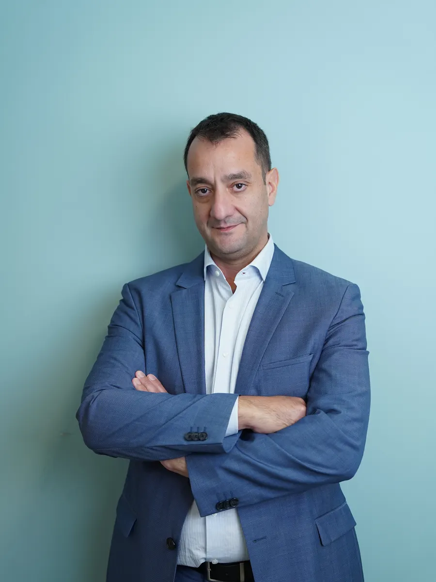 Ο Μάριος Δέτσης με μπλε σακάκι και λευκό πουκάμισο, με σταυρωμένα χέρια, μπροστά σε γαλαζοπράσινο τοίχο