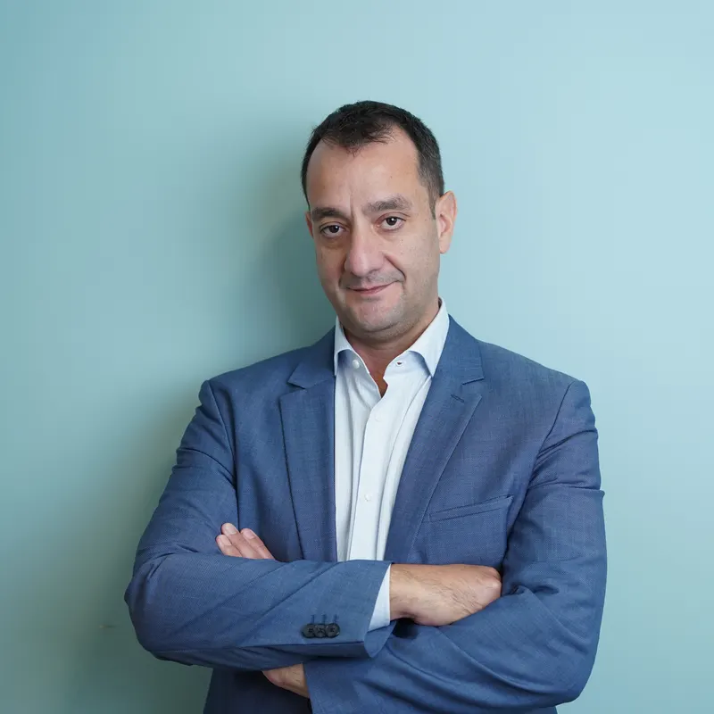 Ο Μάριος Δέτσης με μπλε σακάκι και λευκό πουκάμισο, με σταυρωμένα χέρια, μπροστά σε γαλαζοπράσινο τοίχο